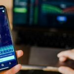 user using a mobile phone and laptop displaying stock market price movements