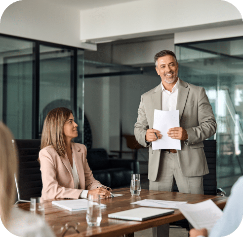 Smiling businessman presenting to team in boardroom with documents in hand